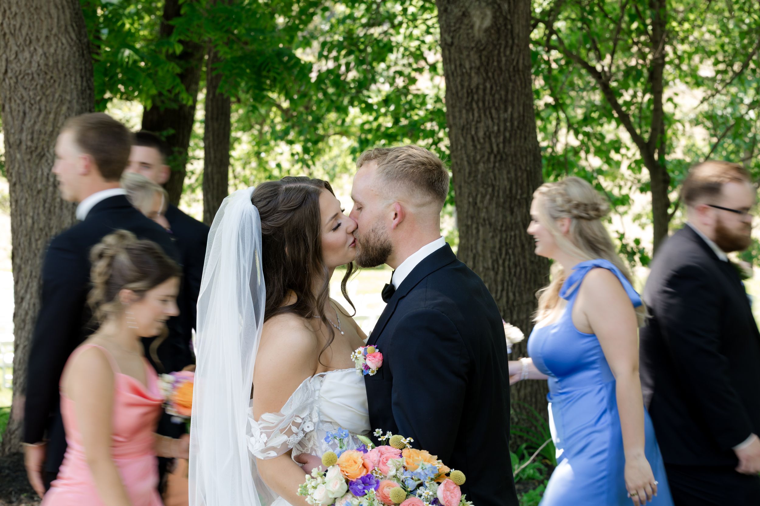 Bride and groom share a kiss outdoors surrounded by wedding party in colorful attire under lush green trees.