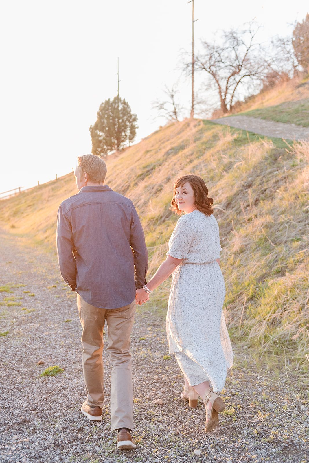 A couple strolls hand in hand along a scenic hillside path at sunset wearing casual attire.