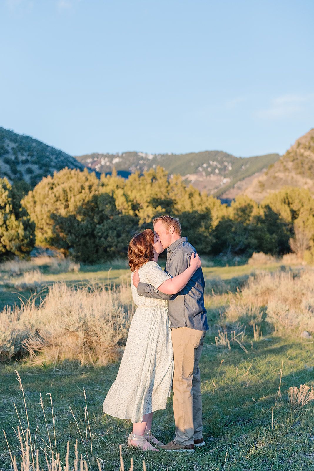 A romantic couple embraces on a mountainside trail during golden hour with mountains in background.