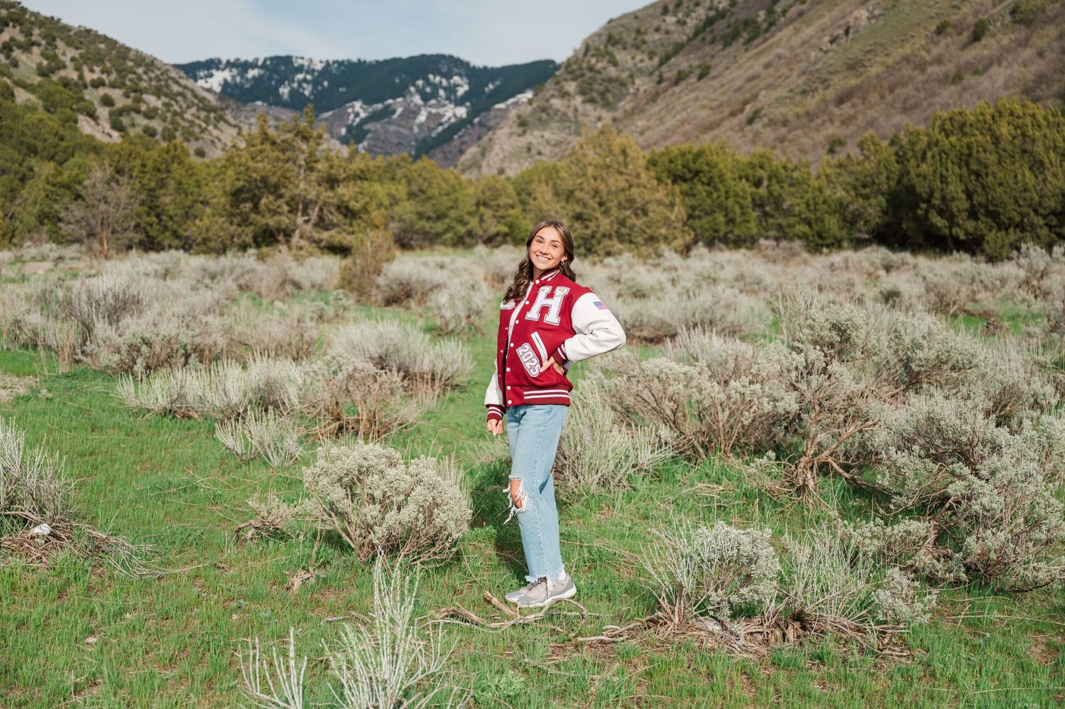 A person wearing a red varsity jacket stands in a mountain meadow surrounded by sagebrush.