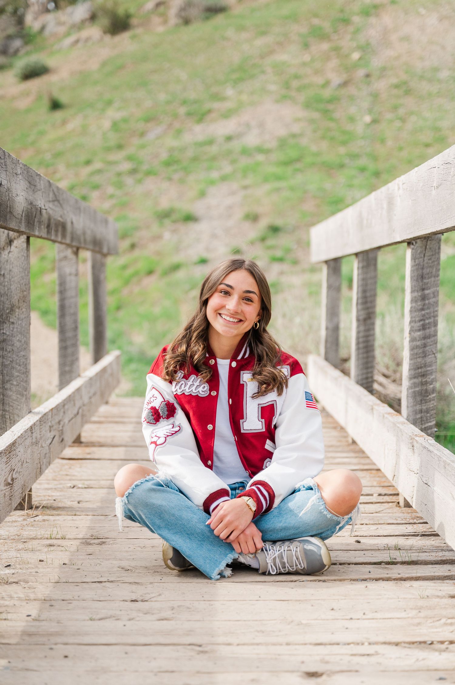 A casual portrait of someone sitting cross-legged on a wooden bridge wearing a varsity jacket.