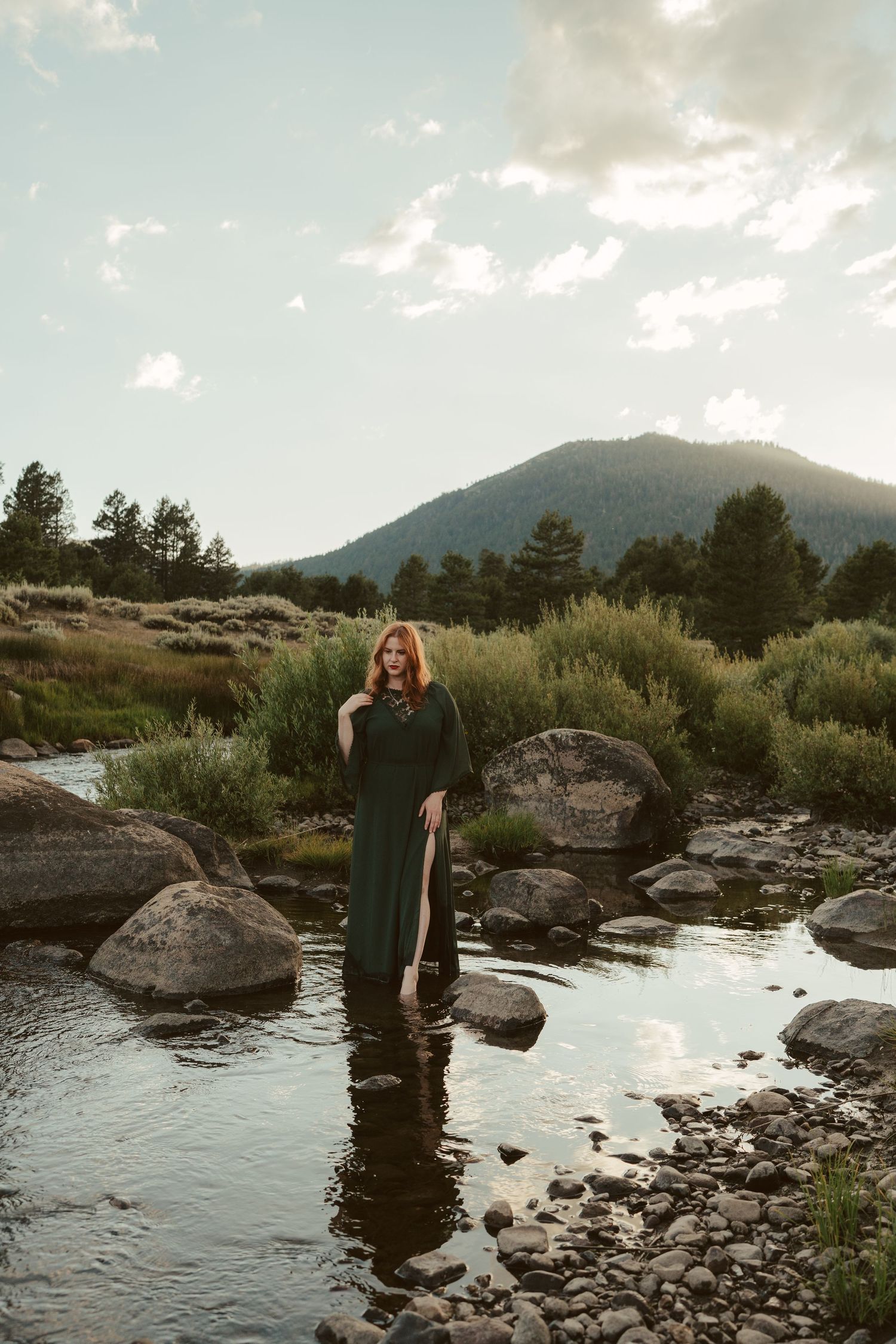 Woman in a green dress standing in a mountain stream at sunset with a scenic mountain backdrop in the background.