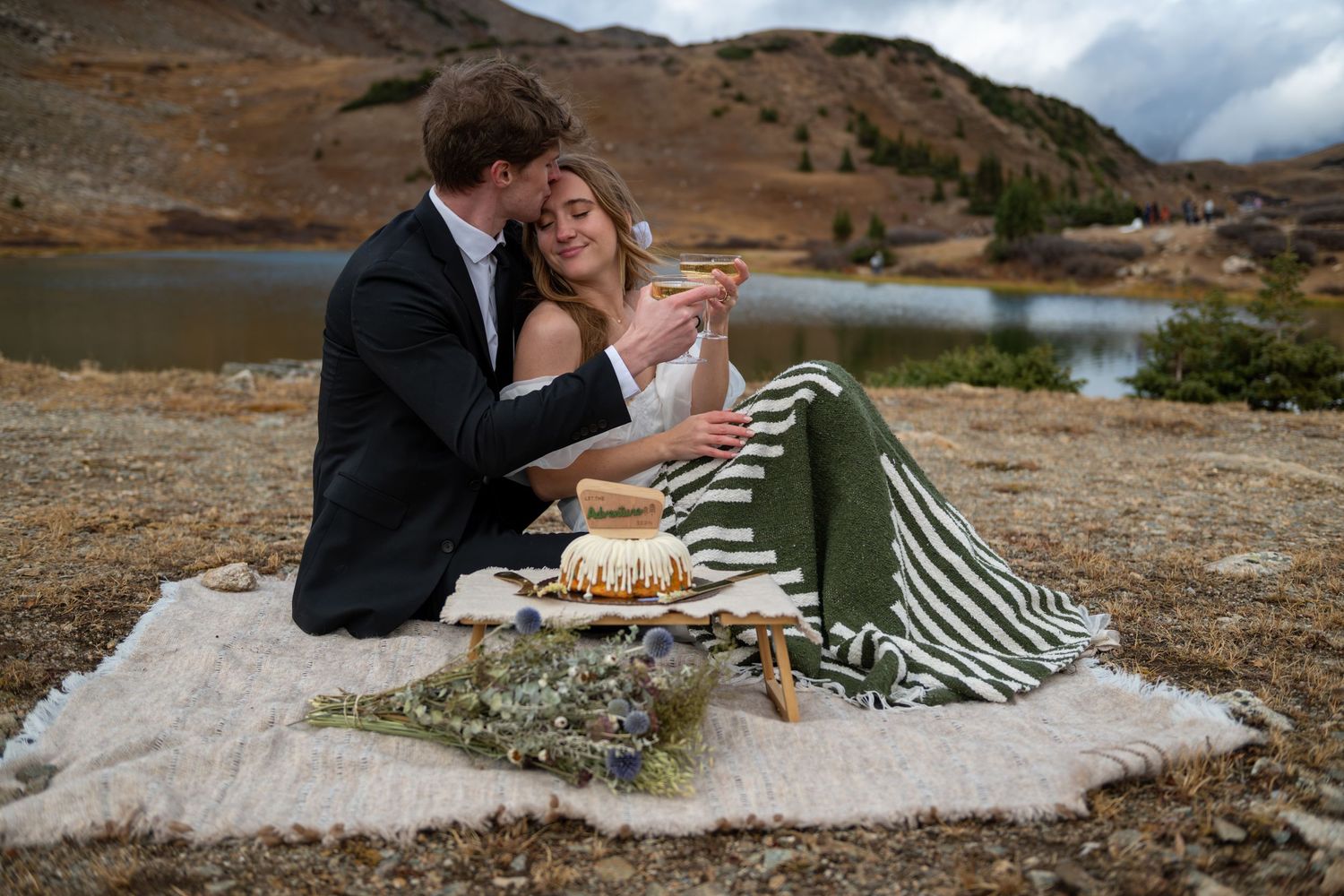 An eloping couple sitting by a lake near Breckenridge, Colorado, sharing wedding cake and champagne with mountain scenery behind them.