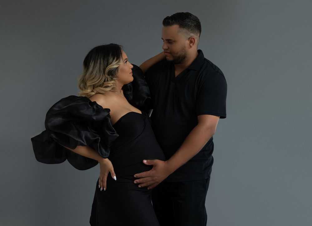 Romantic couple in black attire embrace during maternity photoshoot against grey background.