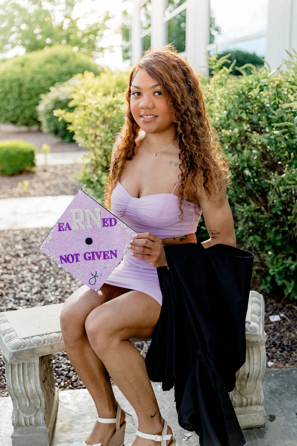 Young woman in lavender dress holding decorated graduation cap reading Earned Not Given, seated outdoors.
