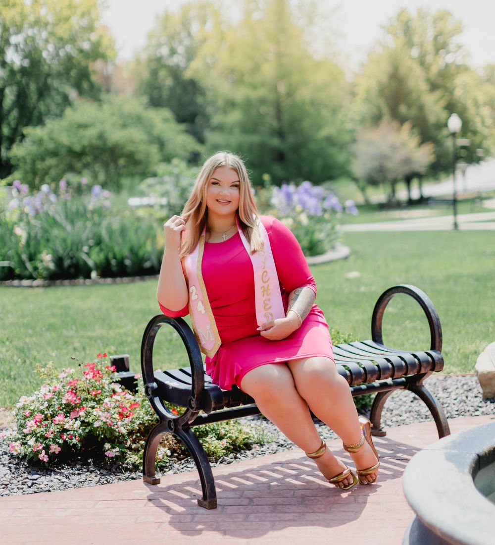 Young blonde woman in pink dress and floral sash poses on park bench surrounded by lush green gardens.