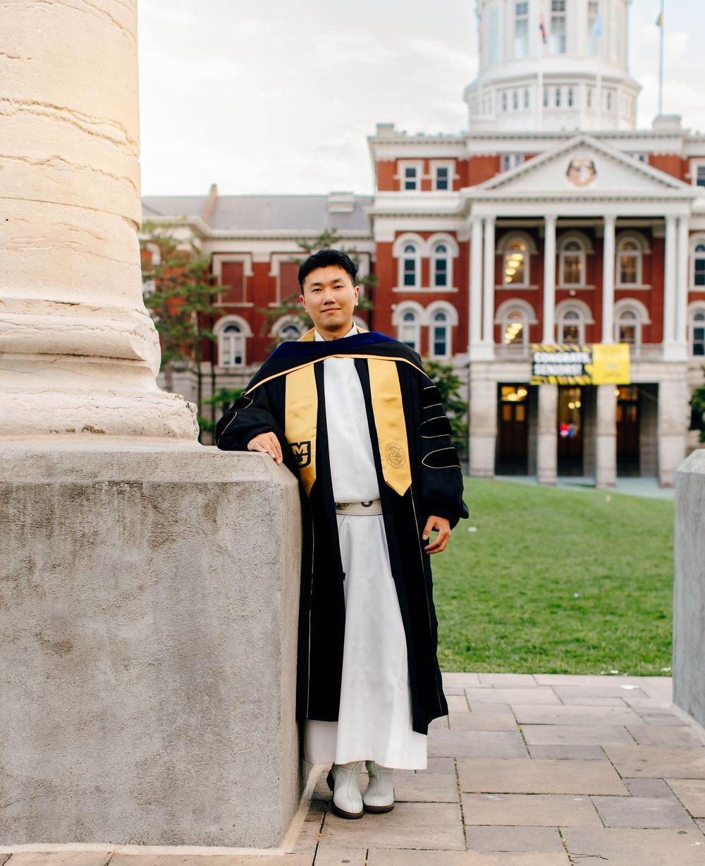 Graduate in doctoral regalia with gold hood poses by stone column before red brick university building.