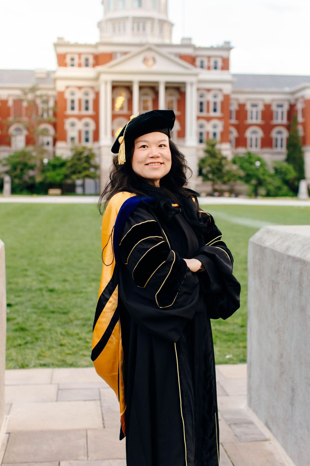 Woman in doctoral graduation gown and cap stands proudly before a red brick university building on a green campus.