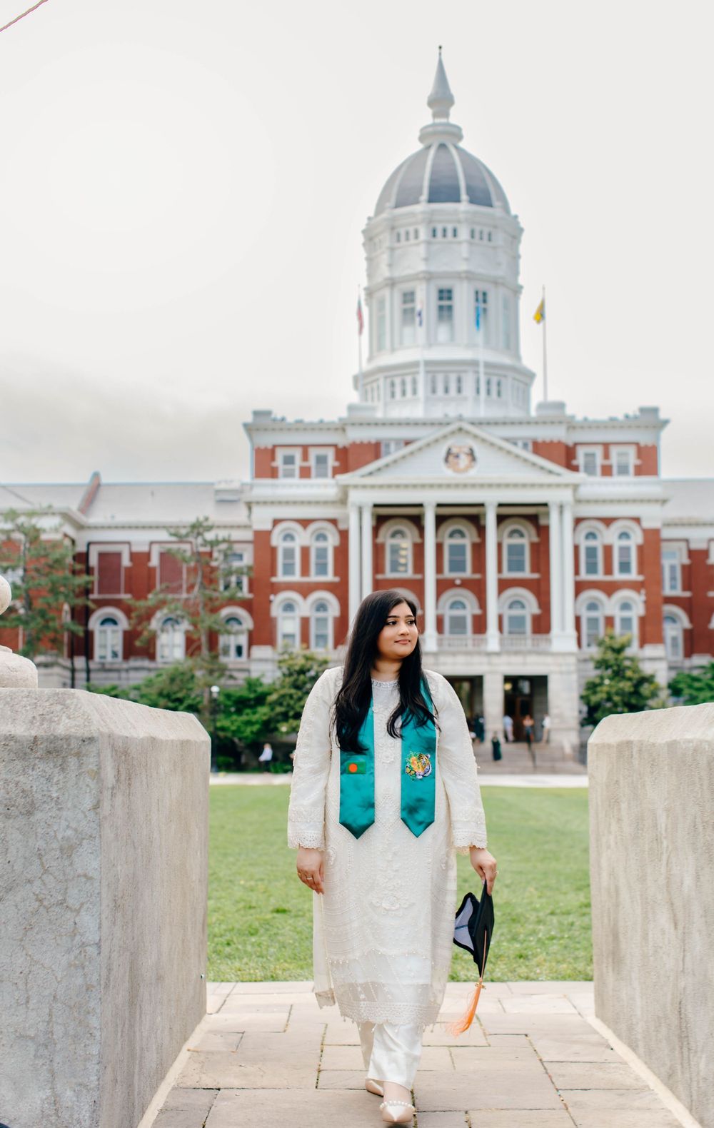 Graduate in white attire and teal stole poses before a grand red brick university building with a dome.