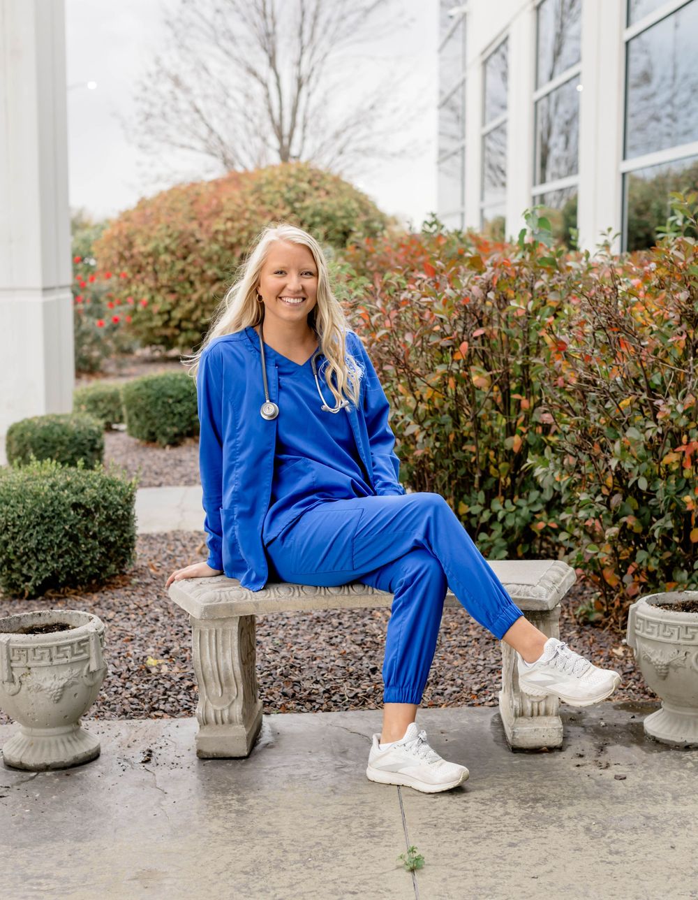 Smiling blonde healthcare professional in royal blue scrubs with stethoscope sitting on stone bench outside medical building.