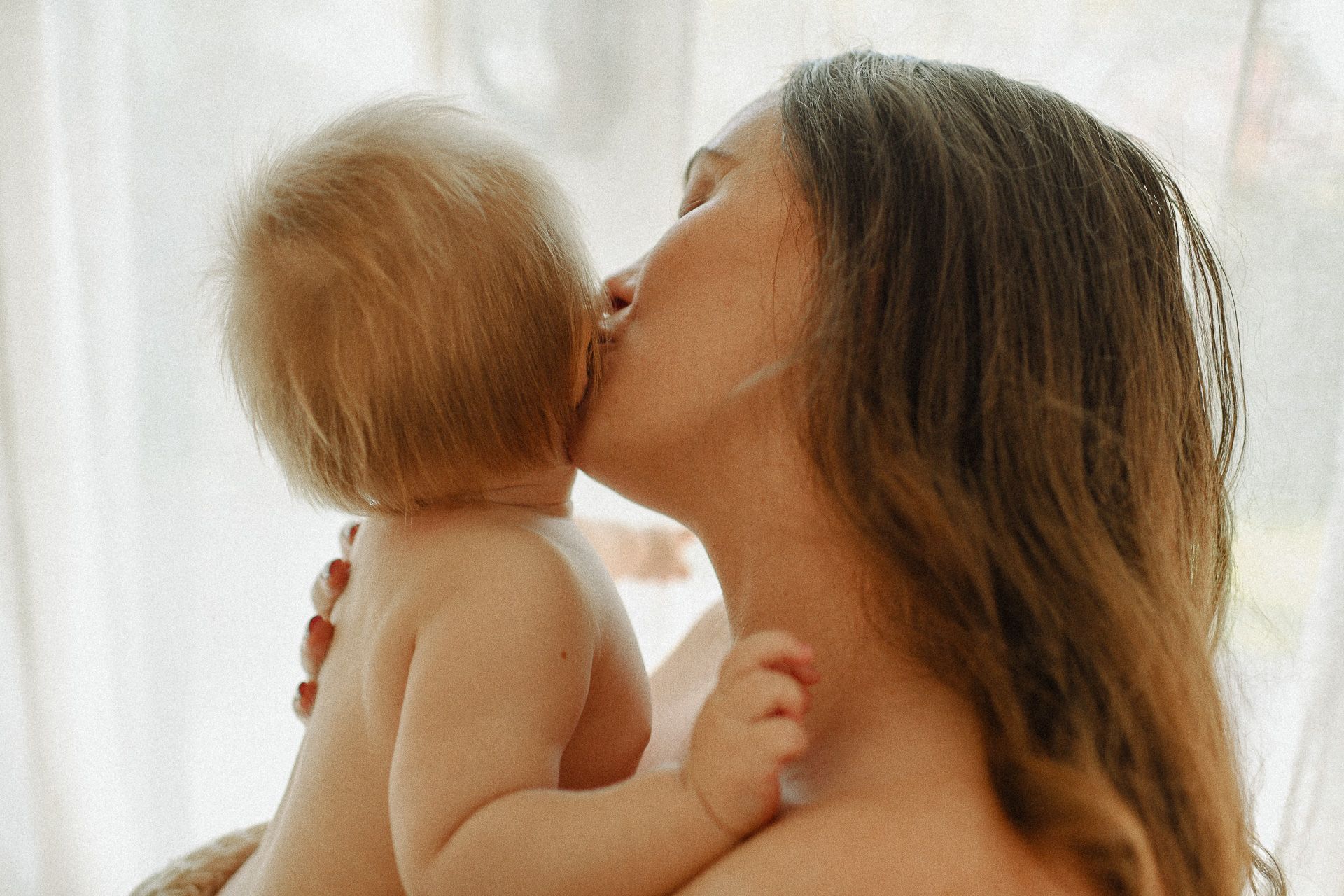 Intimate moment between parent and baby against soft white curtain backdrop.