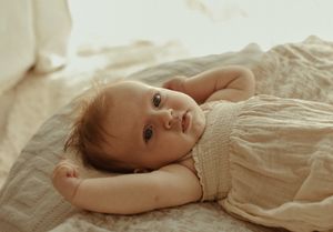Sweet baby in white dress lying on soft bedding in natural window light.