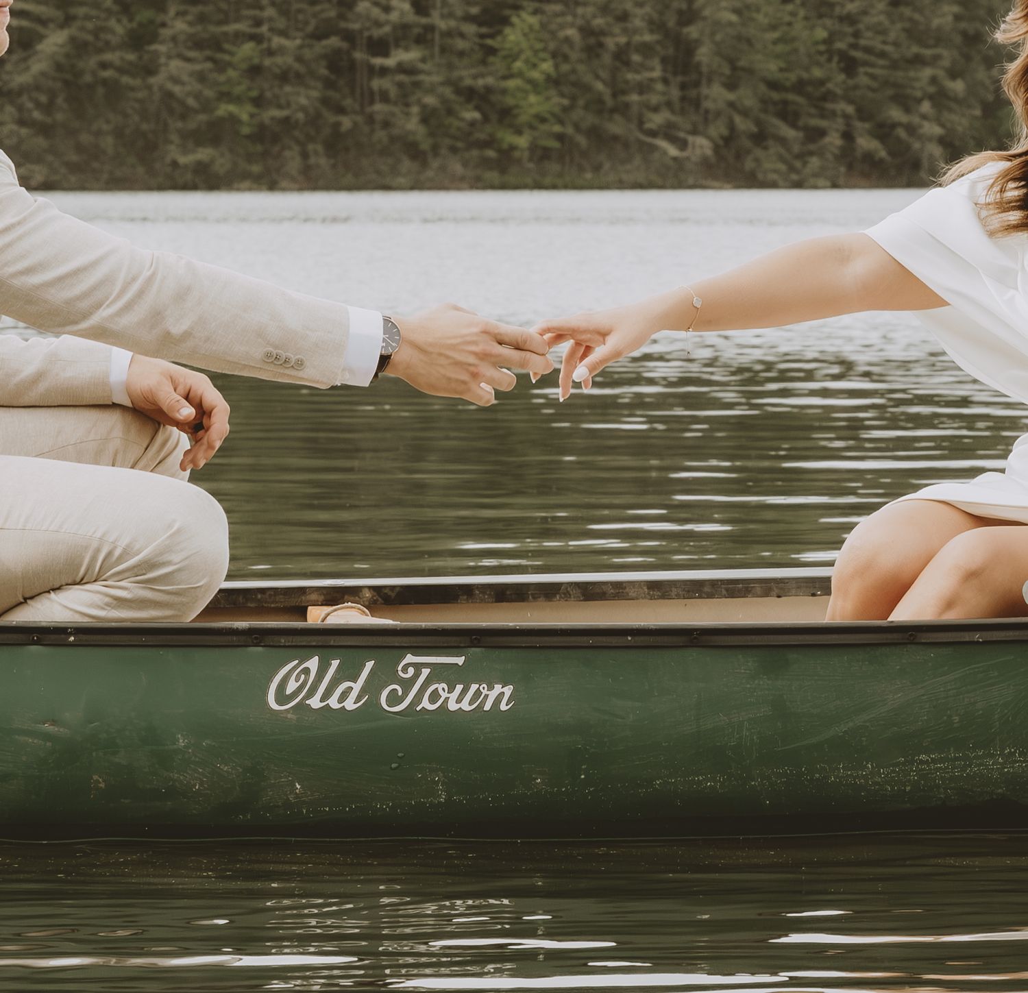 Couple reaching hands across vintage Old Town canoe on calm lake waters.