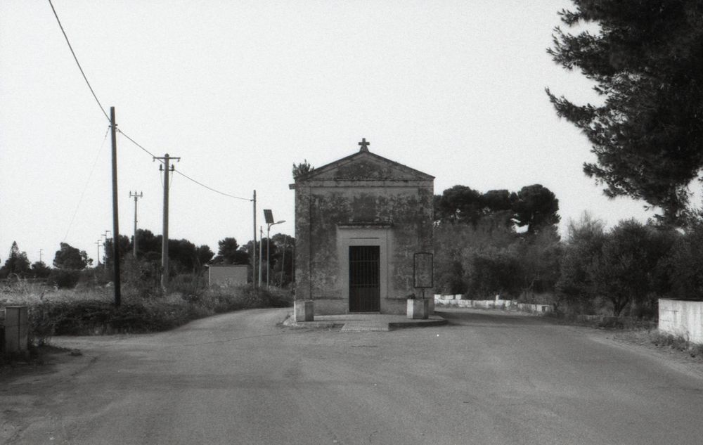 A small stone chapel stands at a rural intersection with utility poles and trees in black and white.