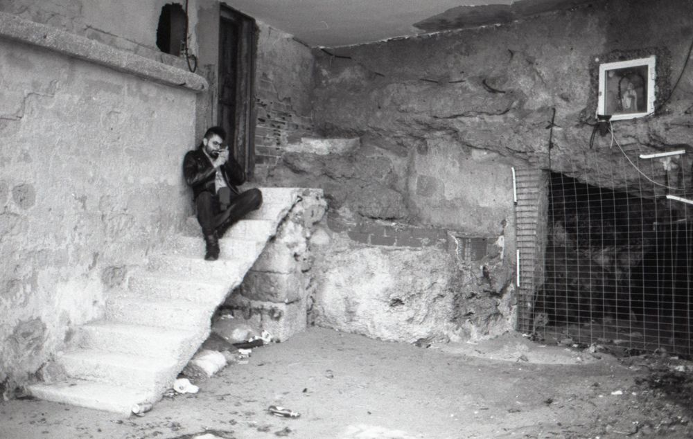 Grainy monochrome image of an underground corridor with stone walls and dim lighting creating an eerie atmosphere.
