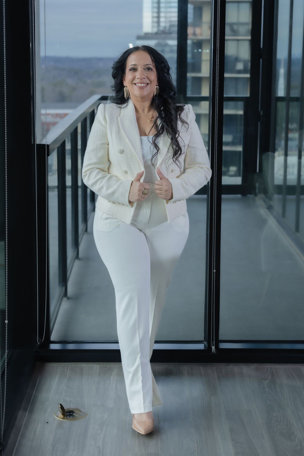 Woman in white blazer poses confidently in modern office hallway with glass windows.