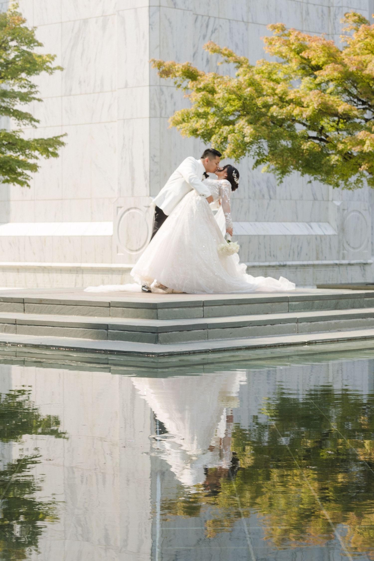 Wedding couple reflected in tranquil pool surrounded by autumn trees and marble architecture.