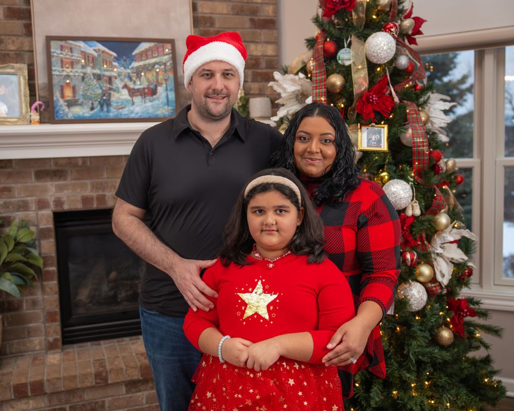 Family of three poses in front of decorated Christmas tree wearing holiday attire with brick fireplace backdrop.