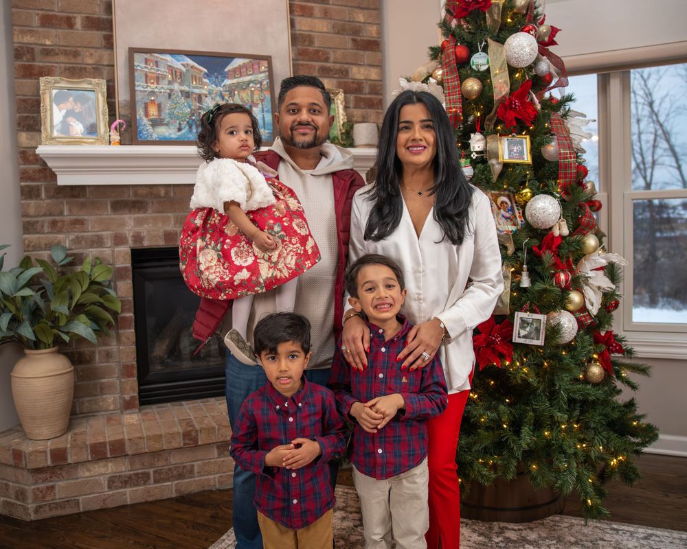 A family poses together by their decorated Christmas tree and brick fireplace in coordinated holiday outfits.