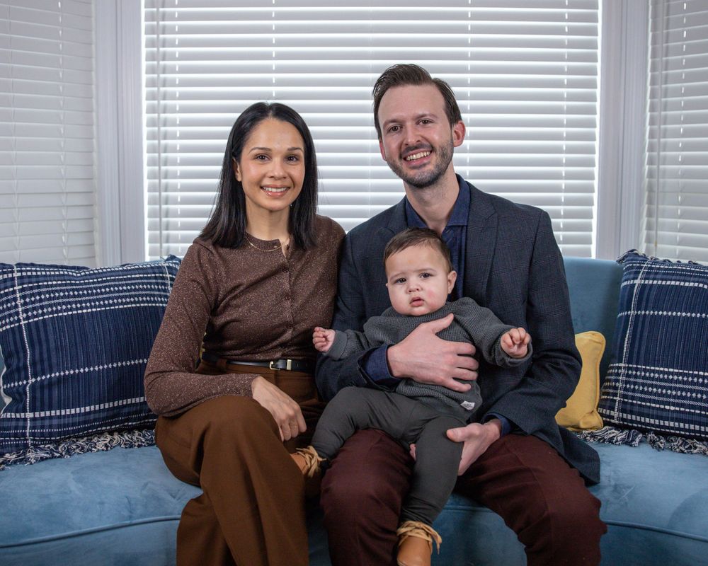 A couple sits on a blue couch with their baby in a cozy home setting with white blinds in the background.