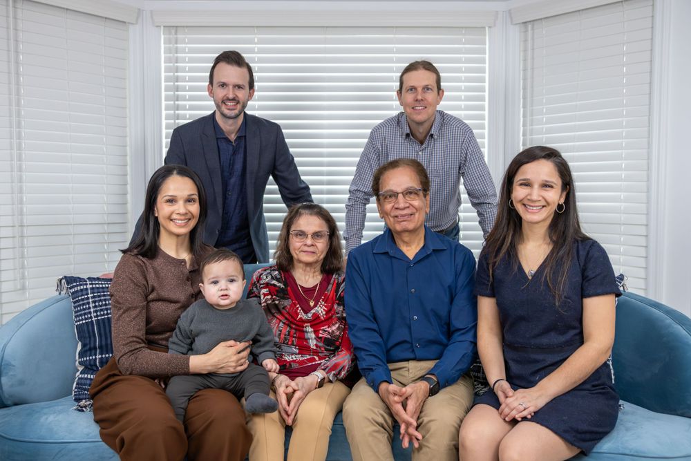 Extended family poses together on blue couch for formal portrait in bright room with white blinds.