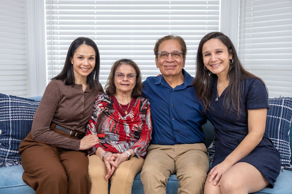 Multi-generational family seated together on blue sofa with white window blinds in background.
