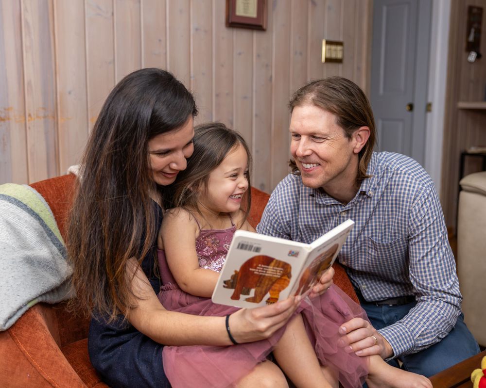 Family reading storybook together on cozy brown couch with wood paneled walls.