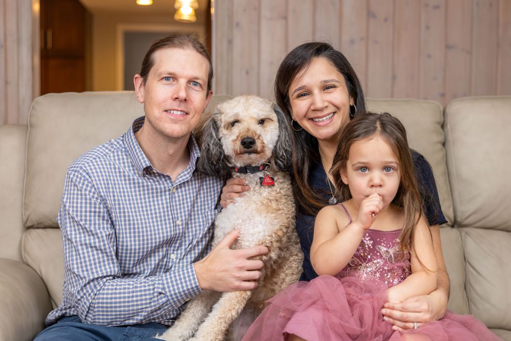 Family portrait with small fluffy dog and young child in pink dress on beige couch.