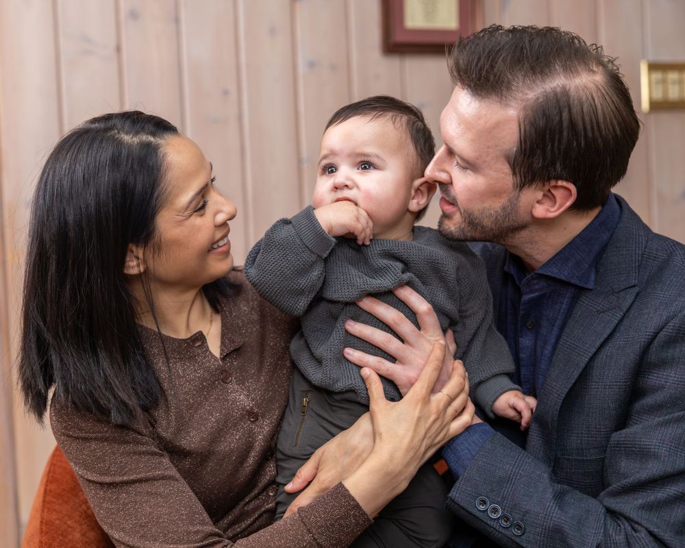 Candid family moment with baby in black outfit against wood paneled wall.