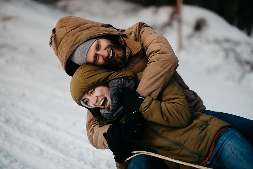 Happy couple in winter jackets embracing and laughing together in snowy landscape.