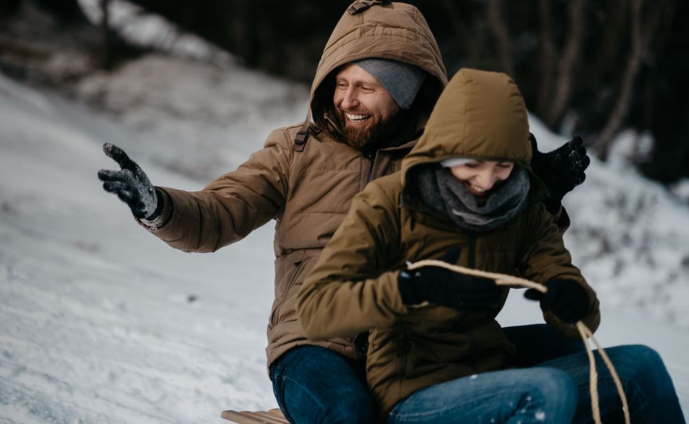 Joyful couple sledding down snowy hill in matching brown winter jackets outdoors.
