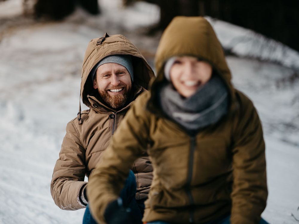 Smiling bearded man and woman in matching brown winter jackets sitting in snow.