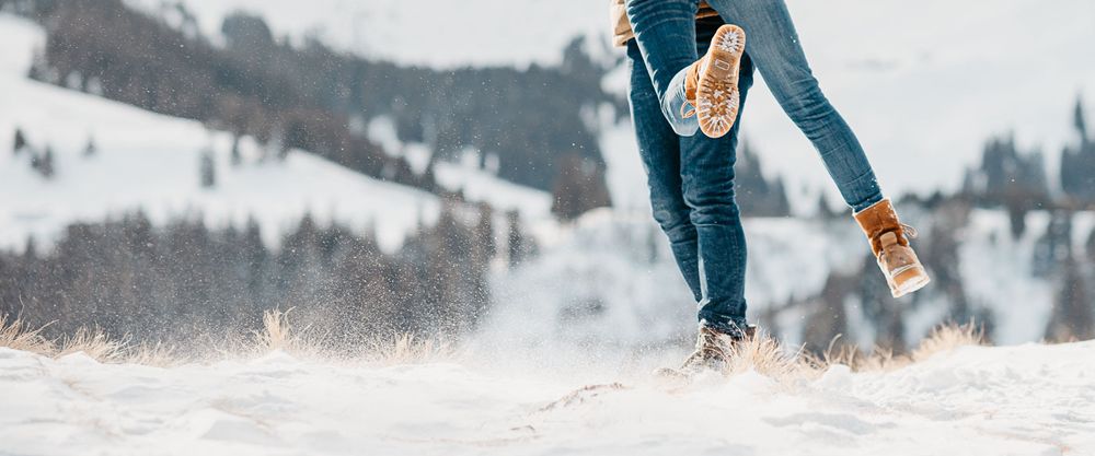 Person walking through snow in jeans and brown boots with snowy hills in background.