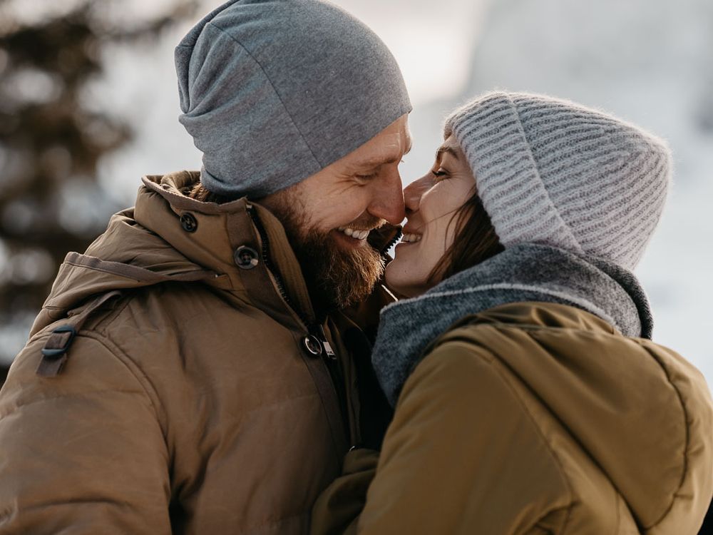 Bearded man and woman in grey beanies sharing a tender moment in snowy winter setting.