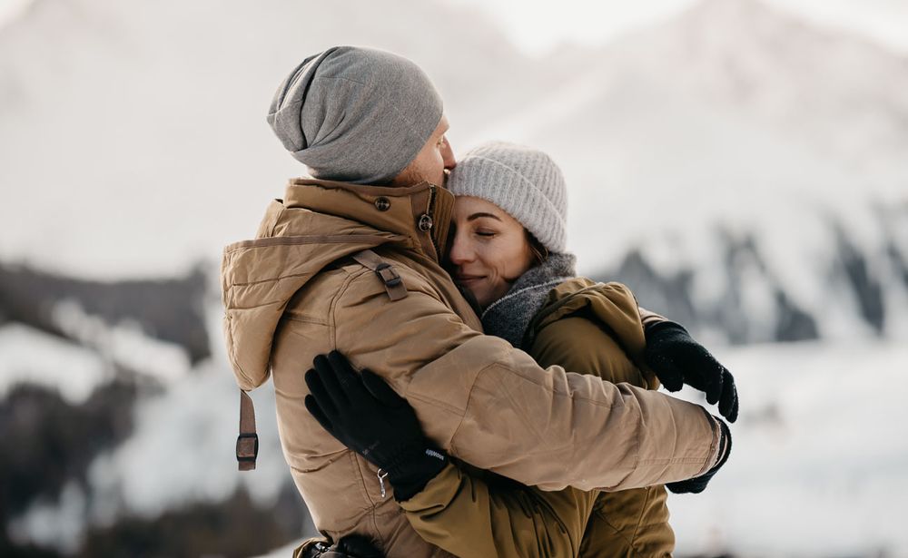 Romantic couple hugging in winter mountains wearing matching beige jackets and grey beanies.