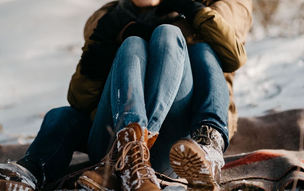 Couple sitting close together in jeans and hiking boots on a blanket in the snow.