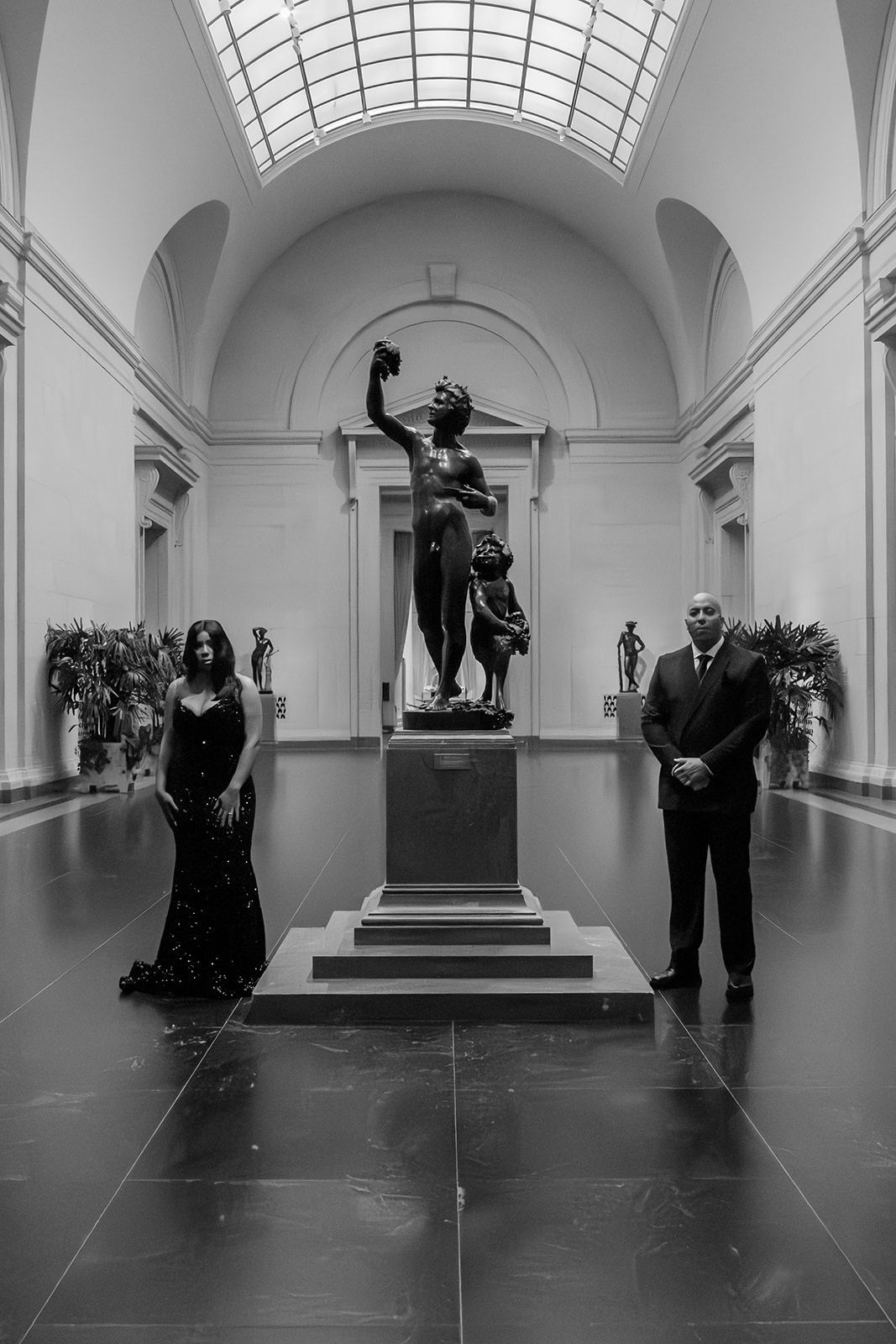 Elegant scene in a museum gallery with an engaged couple posing by a classical bronze statue under vaulted skylight architecture.