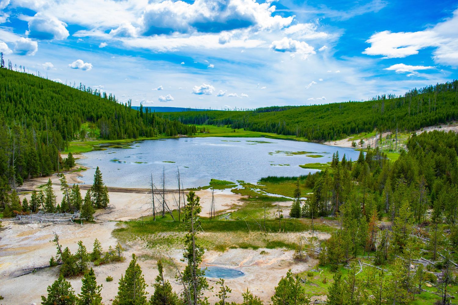 Aerial view of a thermal pool surrounded by green hills and forest in Yellowstone National Park.
