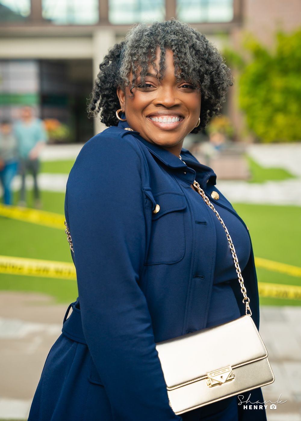 Professional portrait of person in navy blazer with white handbag against outdoor urban background.