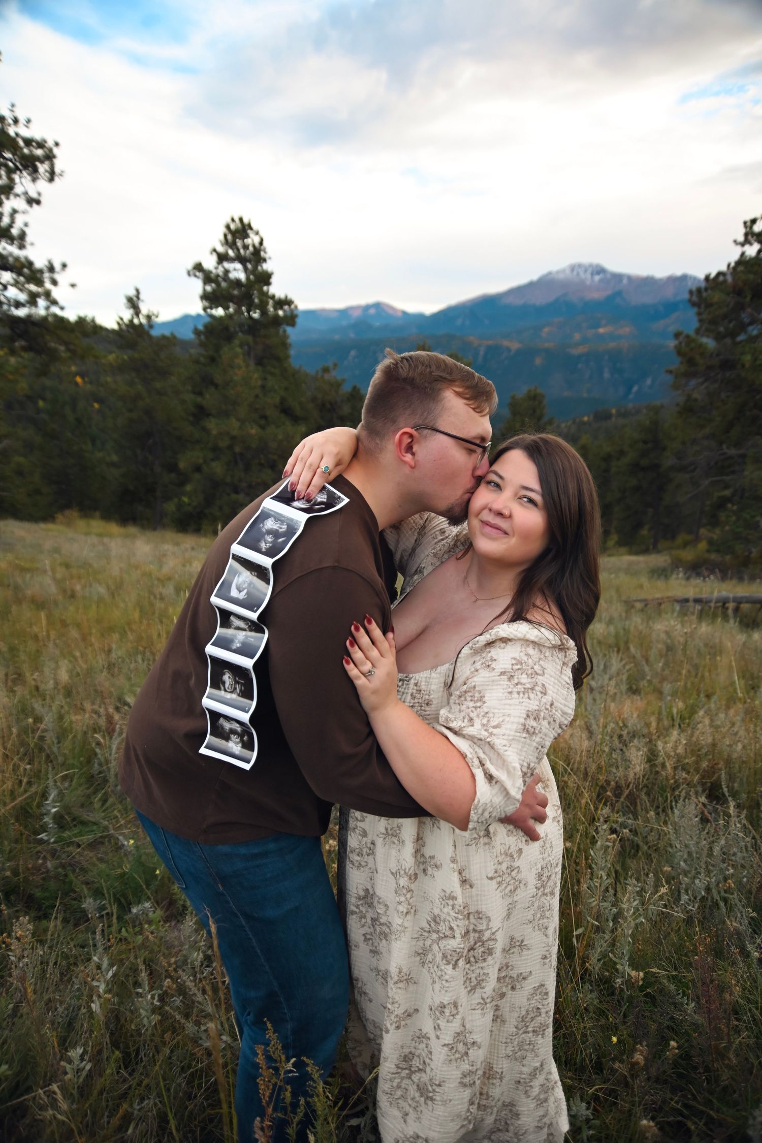 Couple embracing in mountain meadow, man kissing woman's cheek while holding ultrasound photos, announcing pregnancy outdoors.