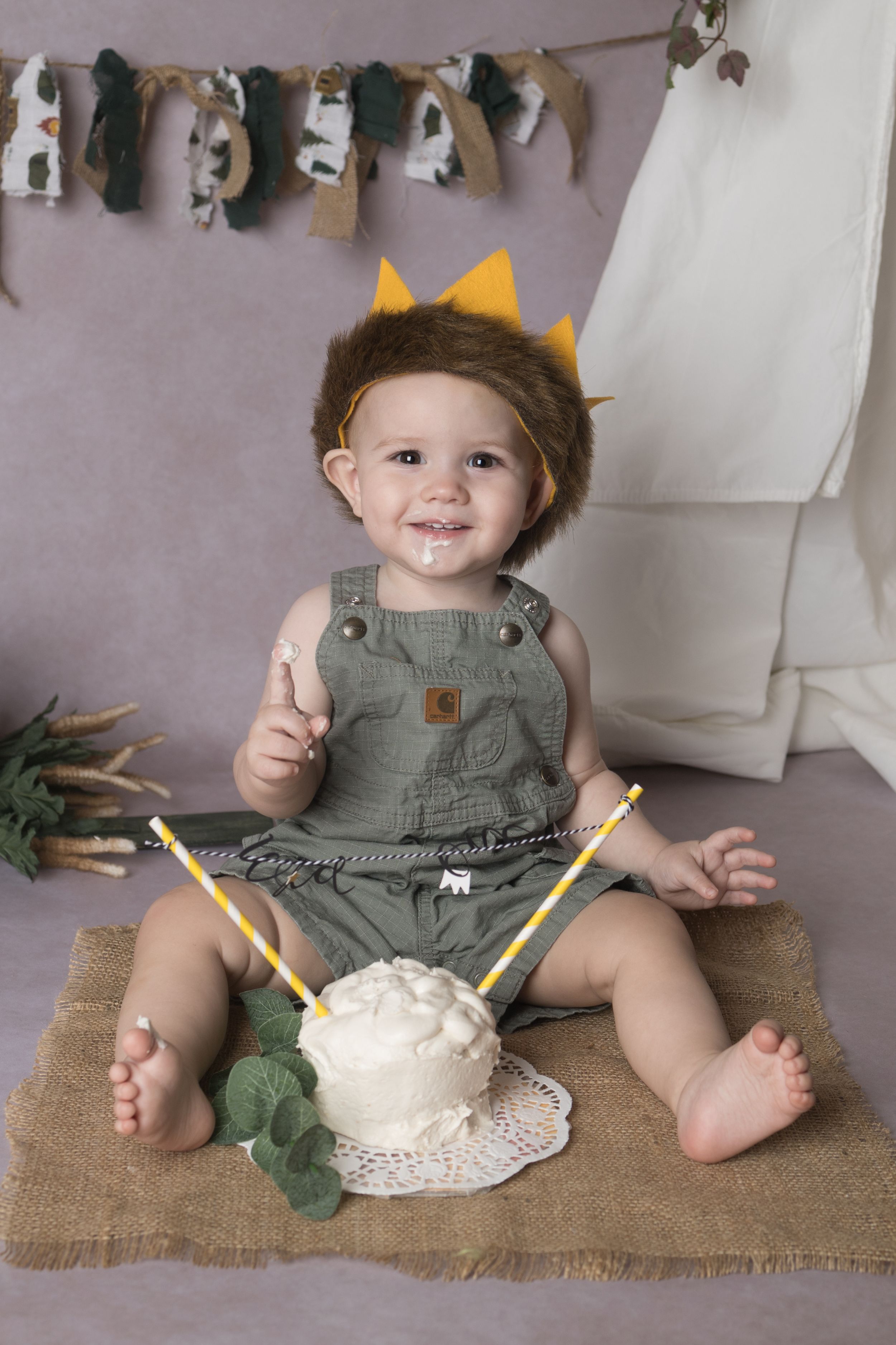 A small child in green overalls and yellow crown sits on burlap with a white cake and decorative bunting overhead.