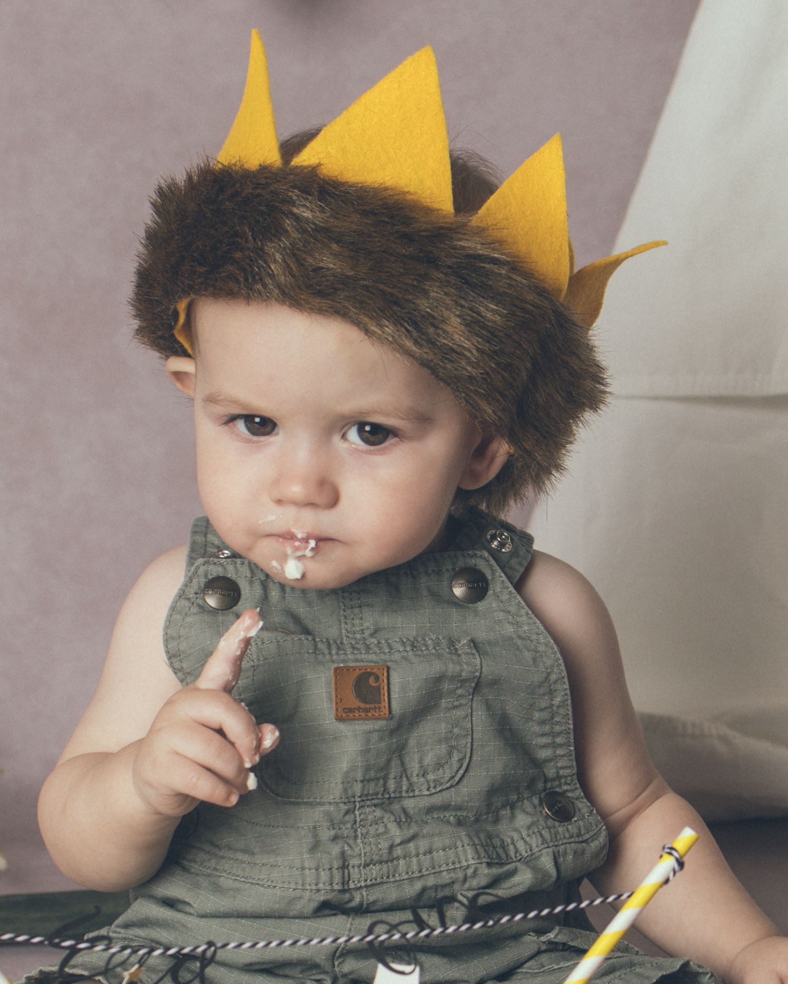 Small child wearing olive green overalls and a yellow felt crown with furry trim sits with a cake on finger.