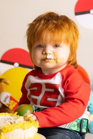 Young child in red jersey with number 22 sits with icing on face and Pokemon-themed party decorations in background.