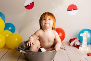 Baby sits in metal basin surrounded by colorful balloons and Pokémon-themed decorations for first birthday celebration.