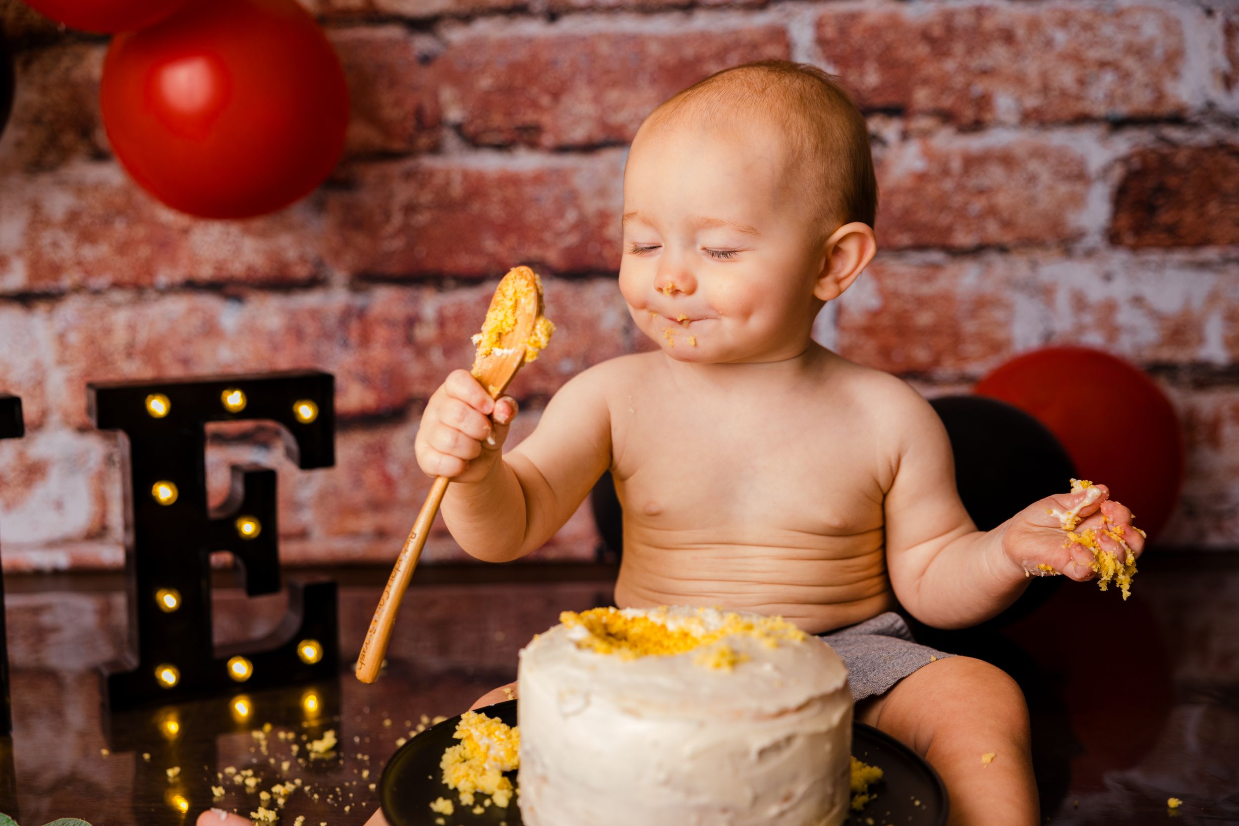 Baby in a high chair enjoying messy cake smash against rustic brick wall background.