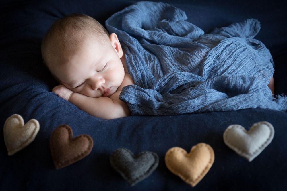 A sleeping baby lies peacefully with decorative heart pillows and soft blue fabric in a newborn photography setting.