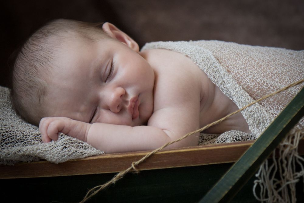 A sleeping baby lies peacefully on white lace fabric with green foliage accents in a dreamy portrait setting.