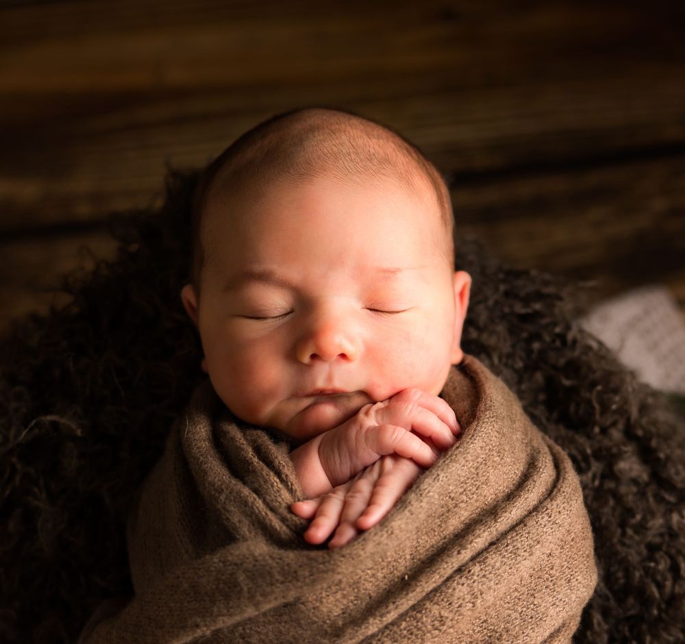 Peaceful sleeping newborn wrapped in soft brown blanket against dark background in professional portrait setting.