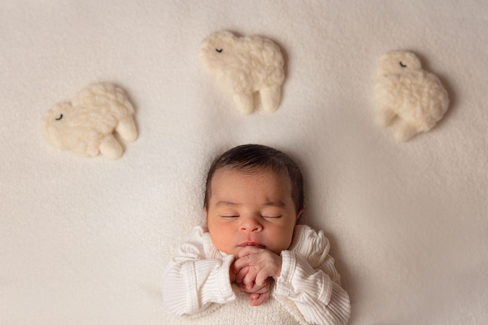 A sleeping infant in white clothing lies peacefully on a cream backdrop with three fluffy white sheep above.