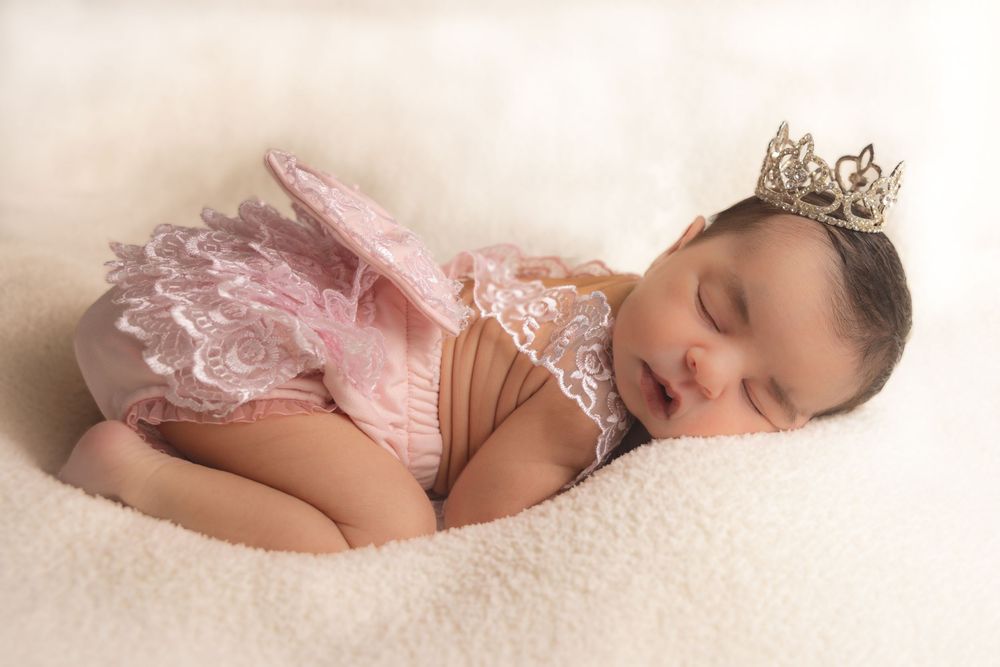Sleeping baby wearing pink tutu and tiny crown rests peacefully on soft white blanket.